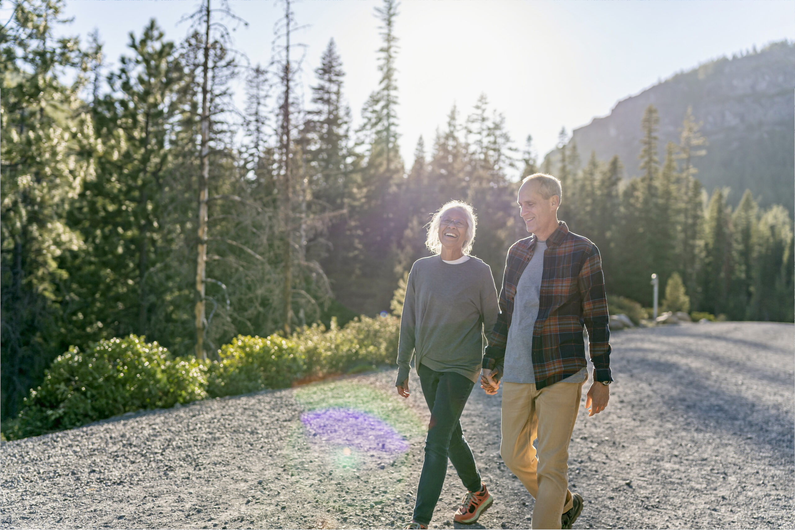 A mixed race senior couple holds hands and talks happily while enjoying a relaxing walk in nature. The happy retired couple is on vacation and hiking in Oregon. It is a beautiful, sunny day and a forest of evergreen trees lines the gravel path they are walking along.